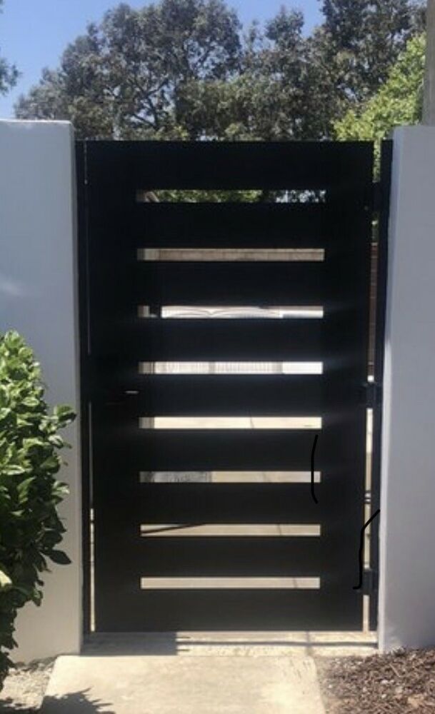 an open black gate in front of a white fence and shrubbery on the sidewalk