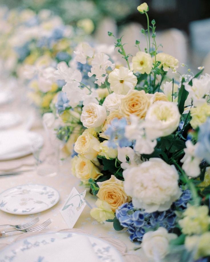 the table is set with white and blue flowers in vases, plates and napkins