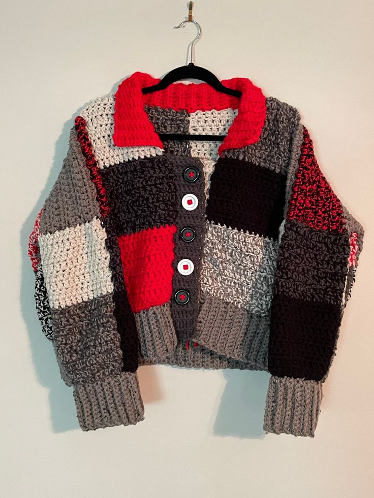 a knitted sweater hanging on a hanger next to a white wall with a red and black button