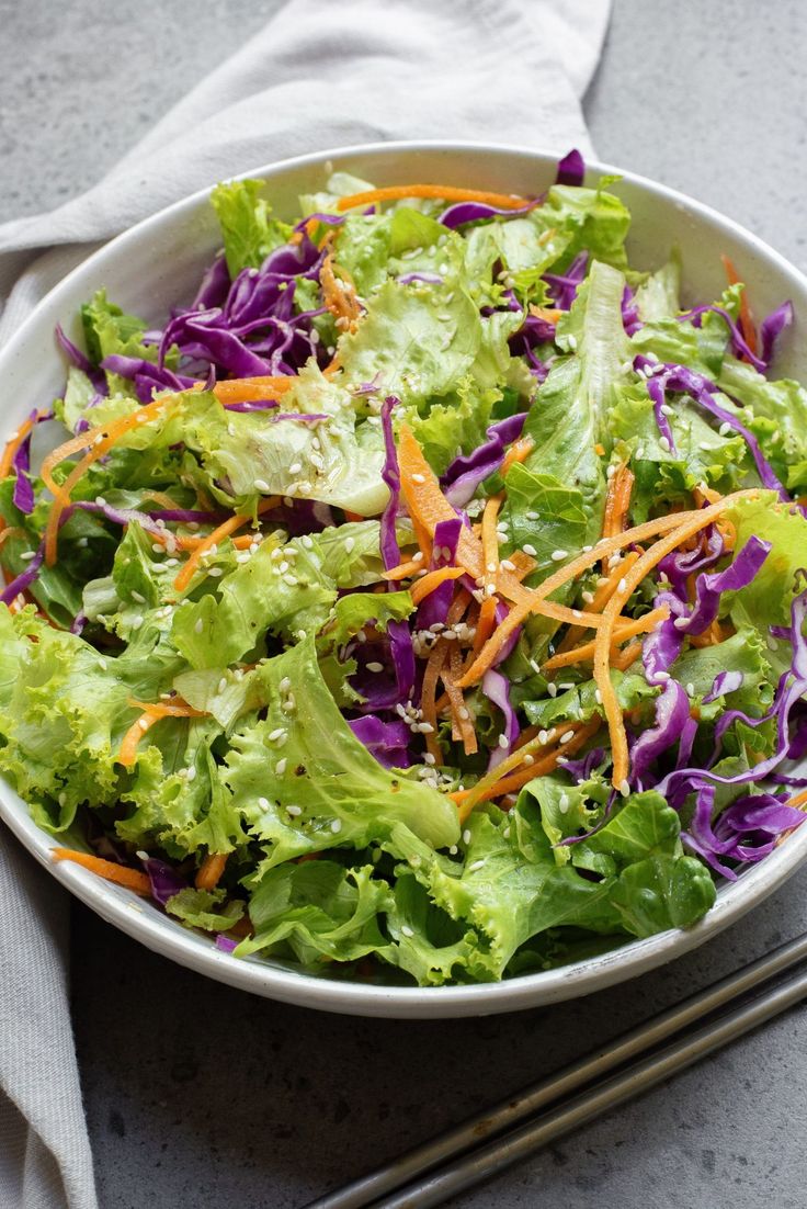 a salad with carrots, cabbage and lettuce in a white bowl next to chopsticks