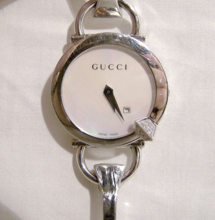 An authentic Gucci ladies Chiodo 122 Diamond Series wrist watch. This is the YA 122505 wristwatch with all of the original packaging and documentation. The wrist-band is 6-3/4" long (measured flat, to the outer of two settings) and the face is 1-3/8" across. It is fully marked on the band, face, back and on all of the packaging - this is a genuine Gucci wristwatch. This watch also includes: -the original brown outer box sleeve &plush-lined box,  -stuffed watch pillow,  -instruction book in its sleeve,  -envelope with two attached strap extenders  the original bar-coded string price tag ($1995.00 CAD).  The watch face is machined mother-of-pearl with excellent colour flash, and it has a date window. The 16 genuine diamonds are of excellent quality and the sapphire crystal is undamaged.  The Modern Jewellery, Old Watches, Watch Box, Women Wrist Watch, Wrist Watches, Modern Jewelry, Silver Watch, Vintage Signs, Mother Of Pearl