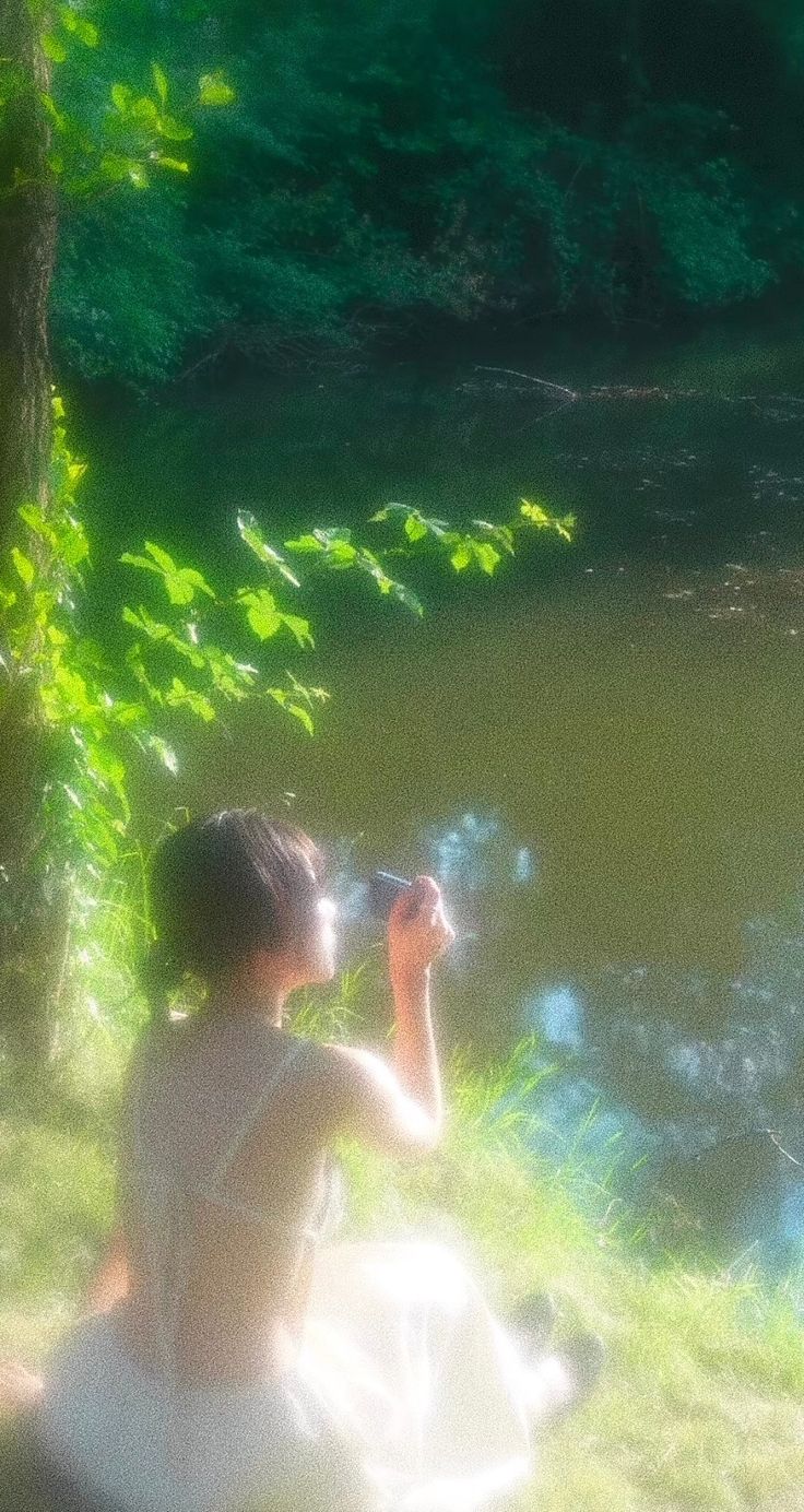 a woman sitting on the ground next to a river drinking from a glass in her hand