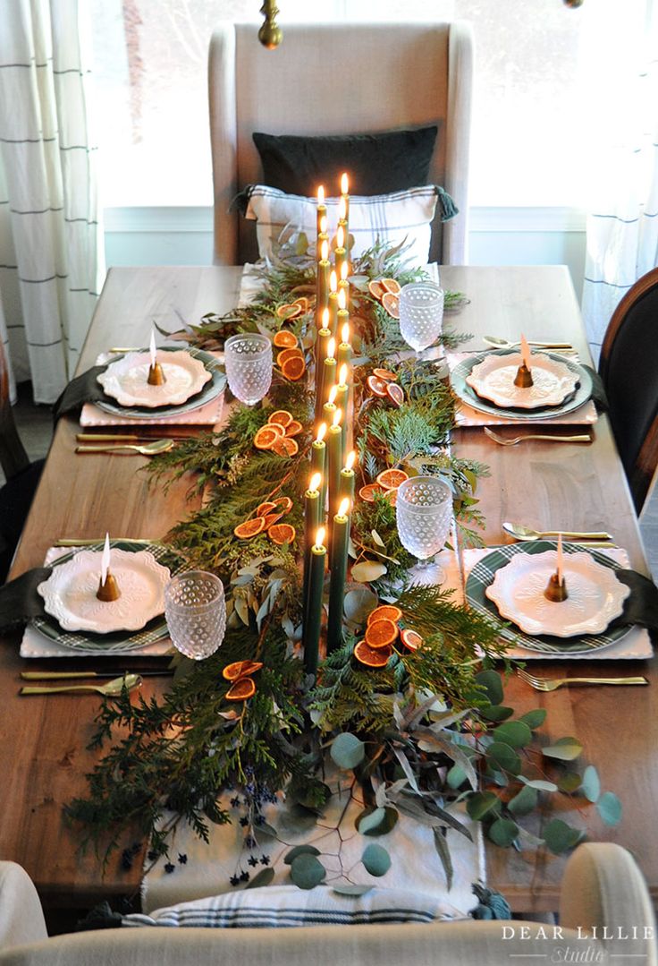 a dining room table set for christmas with candles and greenery on the centerpiece