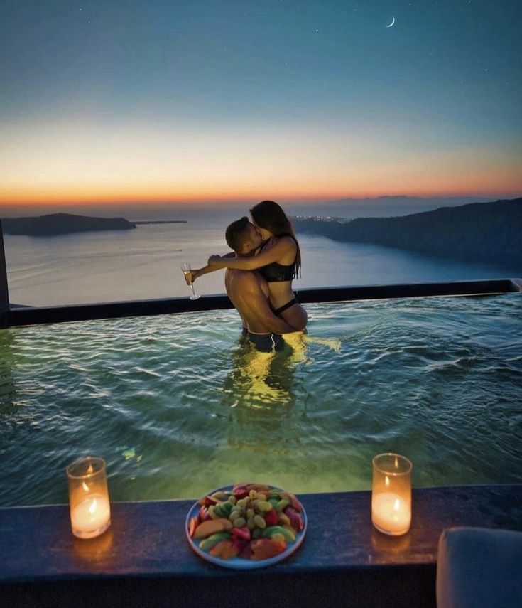 a couple kissing in the water next to candles and plates with fruit on them at sunset