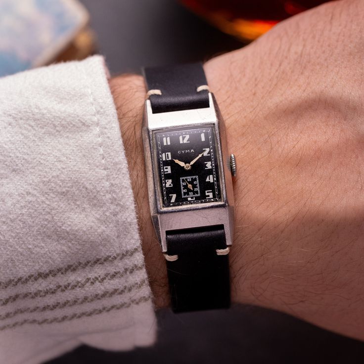 This watch is a vintage Cyma wristwatch from the 1940s, featuring a classic and elegant design indicative of the era. It has a rectangular case measuring 22 x 41 mm, constructed from sturdy stainless steel, which showcases the watch's enduring construction. The face of the watch displays bold Arabic numerals with an art deco influence, giving it a distinct and stylish look. The hands are a simple yet elegant design, complementing the overall aesthetic.  The strap is crafted from high-quality leather, which would have been chosen for its comfort and durability, and it attaches to the watch case with what appears to be original or period-appropriate fixings. The mechanical movement inside the watch is a caliber 335, known for its reliability and craftsmanship, a testament to the Swiss watchm Vintage Rectangular Watch With Subdials, Vintage Rectangular Chronometer Watch, Vintage Analog Watch With Rectangular Dial, Vintage Rectangular Watch Accessories With Diamond Hour Markers, Retro Rectangular Watches For Formal Occasions, Retro Rectangular Formal Watches, Timeless Rectangular Watch Bands With Polished Finish, Art Deco Rectangular Quartz Watch, Vintage Rectangular Analog Watch Accessories