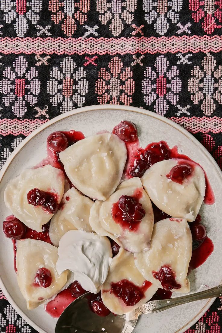 a white plate topped with ravioli covered in cream and cherry sauce next to a spoon