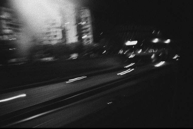 black and white photograph of cars driving down the road at night with buildings in the background