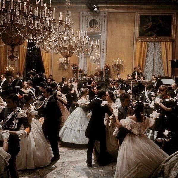 a group of people dressed in formal dress dancing together at a ball with chandeliers hanging from the ceiling