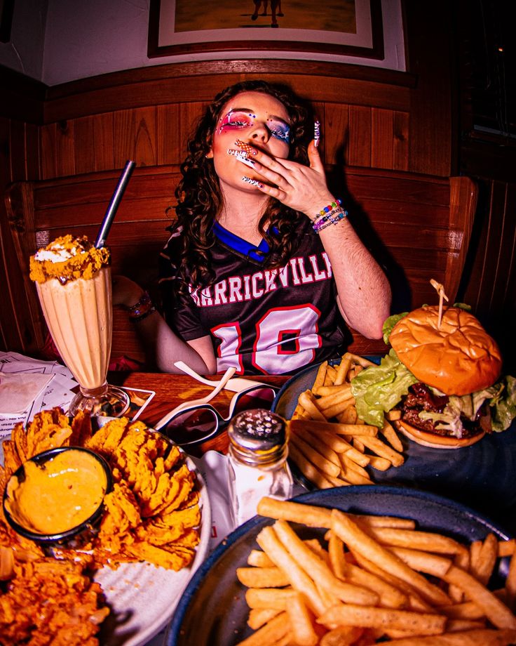 a woman sitting at a table covered in lots of food eating fries and burgers