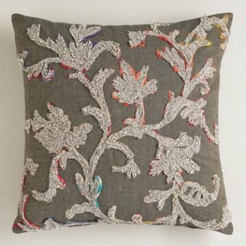 a gray pillow with white and red embroidered flowers on the front, sitting against a wall