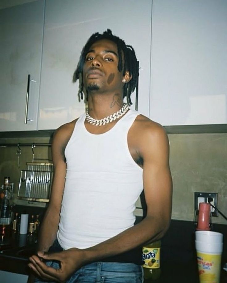 a man with dreadlocks standing in a kitchen wearing a white tank top and blue jeans