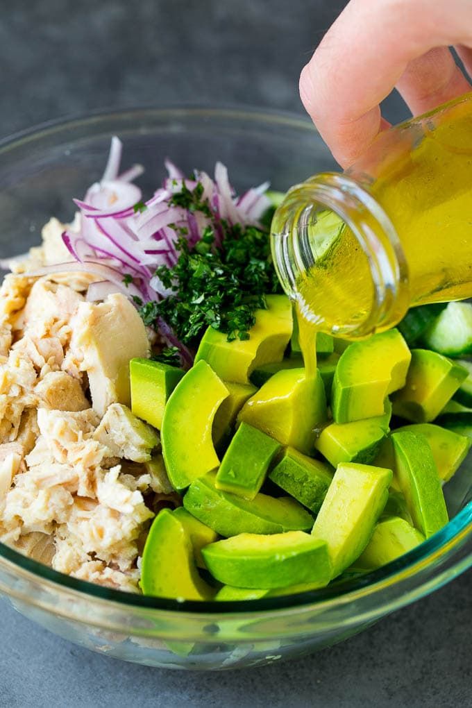 someone pouring dressing into a glass bowl filled with chopped vegetables and chicken, including cucumbers