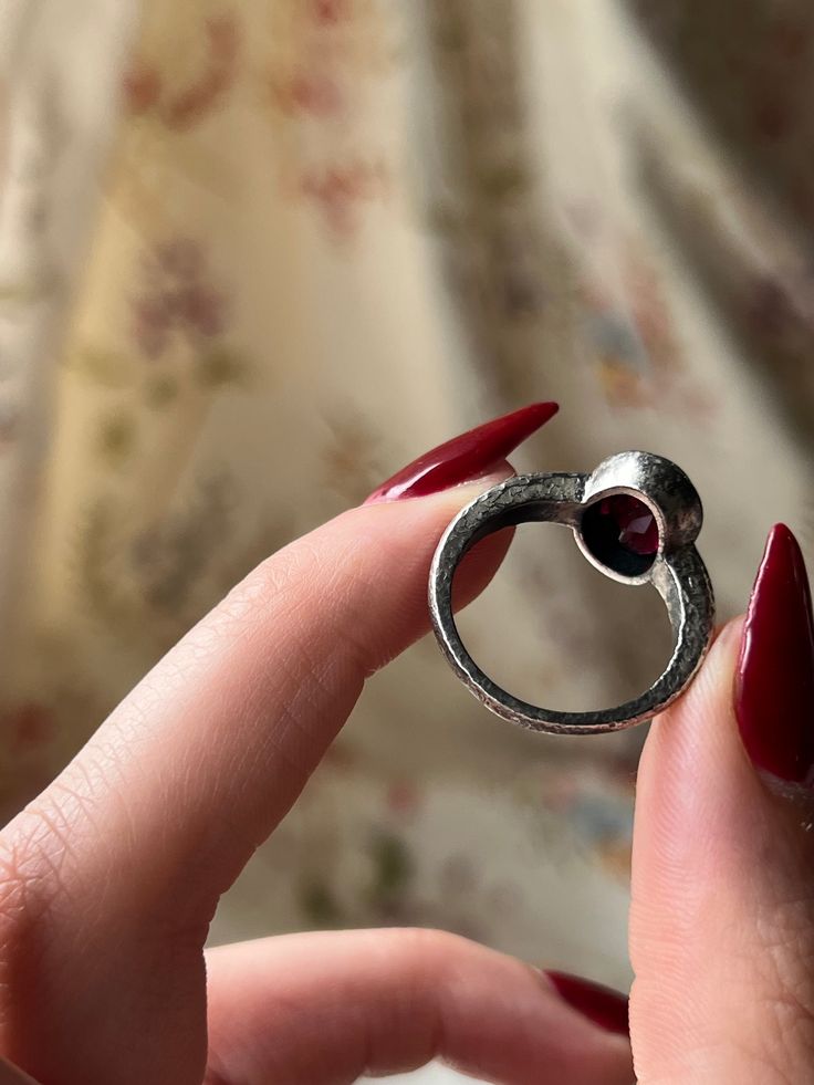 A gorgeous and vampy adornment that is a custom Curio original! These hand-made pieces are a true treasure, for they are one of a kind and crafted with intention to marry the aesthetics of the past, with the modern stylings of the present. This hammered silver garnet ring features a stunning blood-red garnet cabochon set in striking sterling silver. The band is simple and understated, with fine scrolling foliate detail that bring a gentle glamour to the silhouette. There is versatility in this design as well – pair it with a casual or formal look, or even go another step ahead and loop it through a delicate chain as a charm. Either way, this Curio Original is a magnificent addition for any jewelry lover’s collection! Materials and Features: Sterling Silver, Garnet Cabochon Age/Origin: Curi Vintage Hand Forged Jewelry For Anniversary, Heirloom One-of-a-kind Jewelry For Anniversary, Hand Forged Ruby Ring Jewelry, Hand Forged Ruby Ring, Hand Forged Vintage Jewelry, Hand Forged Vintage Promise Jewelry, Hand Forged Ruby Ring For Anniversary, Vintage Hand Forged Round Jewelry, Spiritual Hand Forged Rings For Anniversary