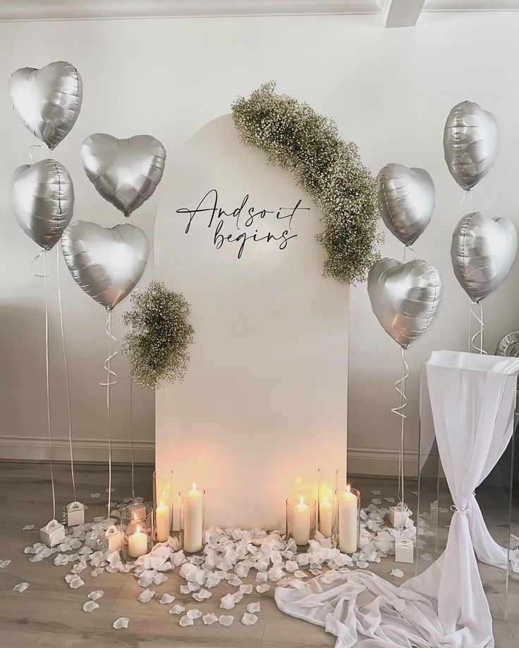 a white backdrop with silver balloons and greenery on the side, surrounded by candles