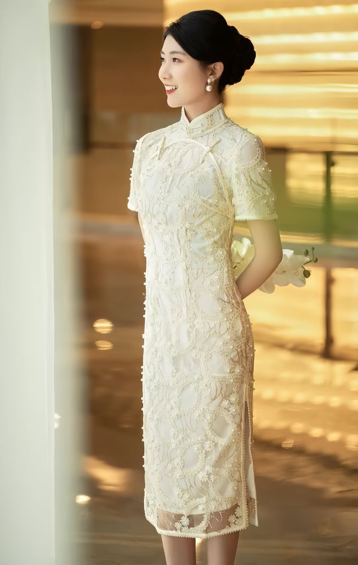 Introducing the stunning short sleeves white lace qipao with delicate floral appliques and faux pearls, a true masterpiece in traditional Chinese fashion. This gorgeous dress is crafted from high-quality lace material that feels soft and comfortable against the skin.The dress features intricate floral appliques that are intricately sewn onto the fabric, creating a beautiful and intricate design that is both feminine and elegant. Faux pearls are also incorporated into the design, adding a touch o Elegant Embroidered Cheongsam For Spring, Elegant White Cheongsam For Spring, Traditional White Cheongsam With Stand Collar, White Fitted Cheongsam With Stand Collar, Elegant Ao Dai With Intricate Embroidery For Ceremony, Elegant Embroidered Cheongsam With Stand Collar, White Cheongsam With Short Sleeves, Elegant White Ao Dai For Formal Occasions, Elegant Floral Embroidered Dress With Stand Collar