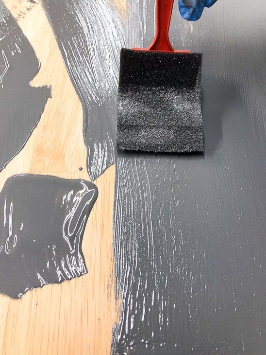 a person using a brush to paint the floor with black paint on it and another hand holding a red tool