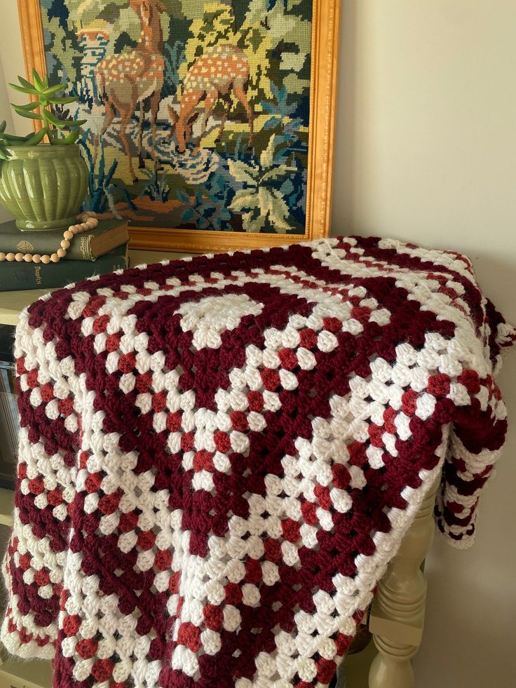 a red and white crocheted blanket sitting on top of a wooden table next to a painting