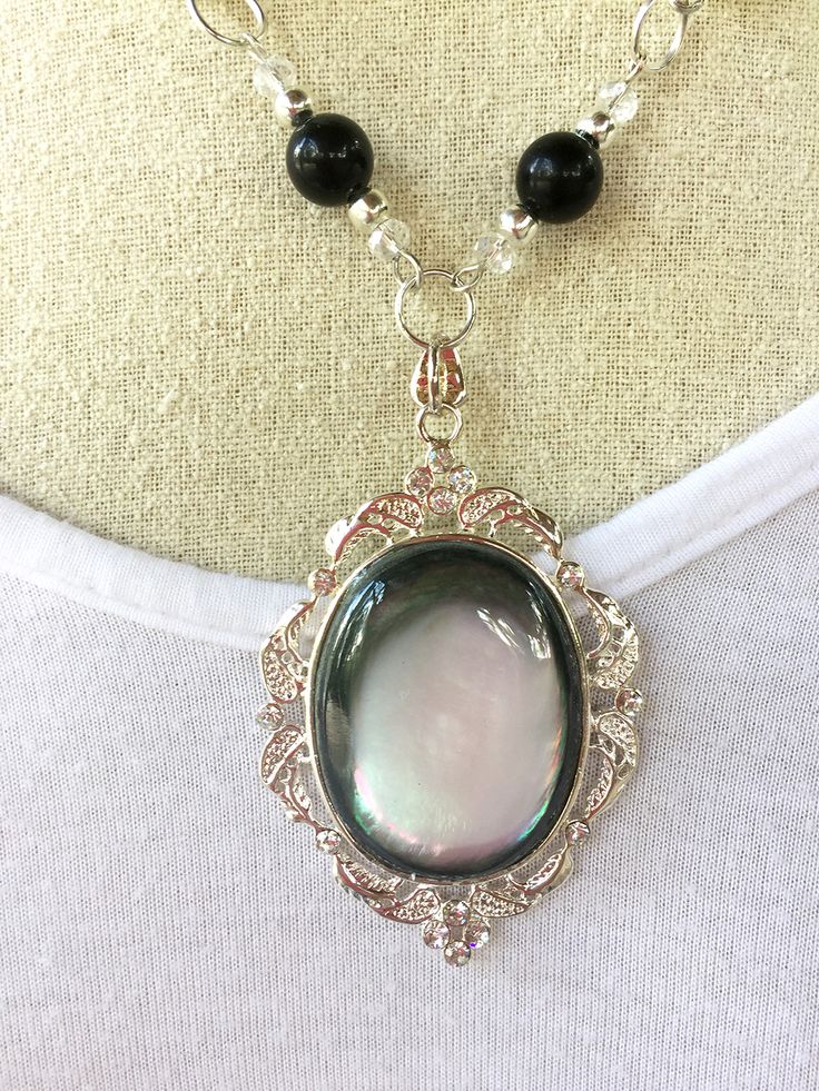 Links of black and white glass rounds, silver tone spacers and clear faceted glass rondelles are added to this filigree opalescent oval pendant. Silver Teardrop Czech Glass Jewelry, Elegant Glass Jewelry With Cabochon, Elegant Iridescent Beaded Jewelry, Elegant Oval Glass Jewelry, Silver Glass Cabochon Necklaces, Silver Glass Cabochon Necklace, Silver Necklace With Cabochon Glass, Elegant Silver Jewelry With Glass Material, Elegant Silver Jewelry With Glass