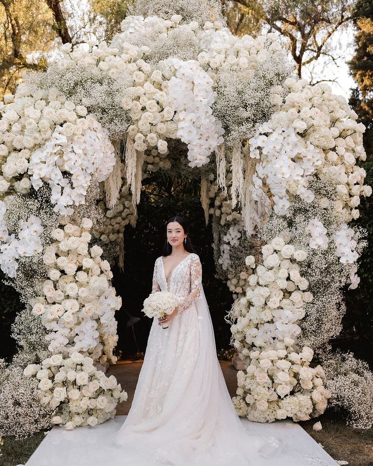 a woman in a wedding dress standing under an arch of white flowers and greenery