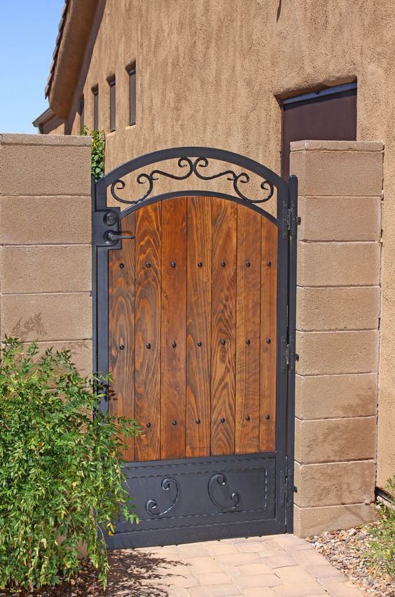 an iron gate with wooden doors in front of a stucco wall and green shrubbery