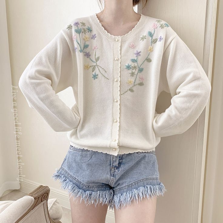 a woman wearing shorts and a sweater with embroidered flowers on it's sleeves, standing in front of a white background