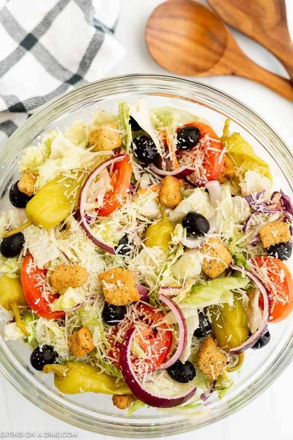 a salad in a glass bowl with olives, tomatoes and croutons on the side