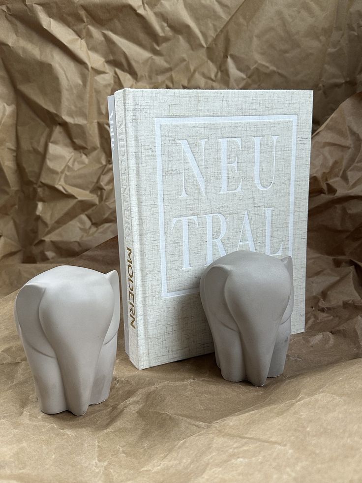 two ceramic elephants next to a book on a brown paper covered surface with the words neu tral written in white