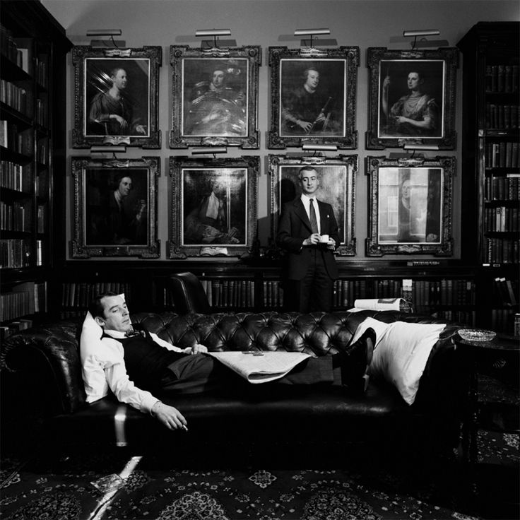 an old black and white photo of two men sitting on a couch in front of bookshelves