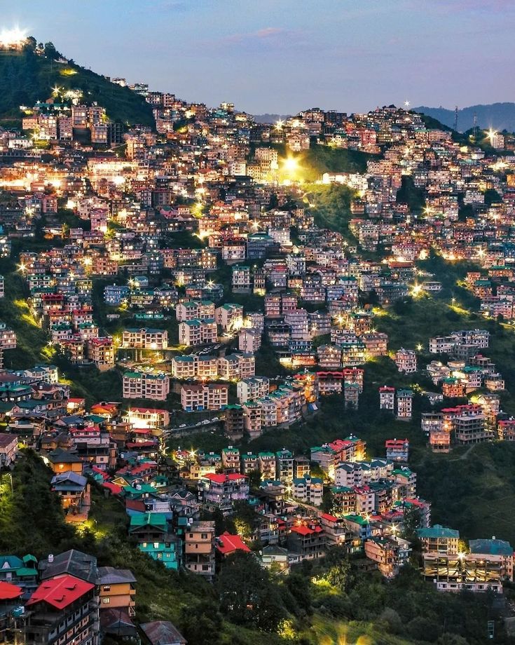 the city is lit up at night with lights on it's buildings and mountains in the background