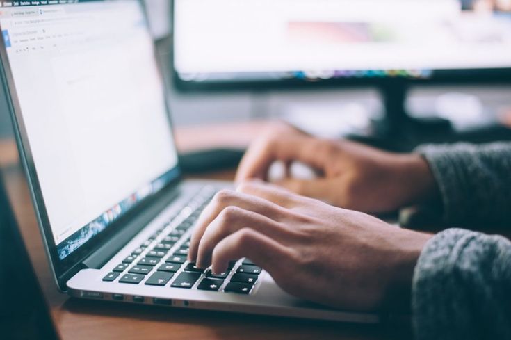 a person typing on a laptop computer at a desk with two monitors in the background