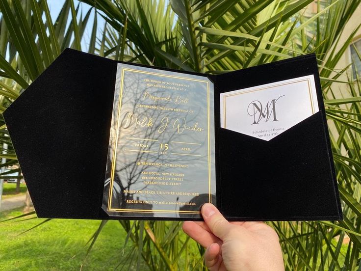 a person holding up an open black and gold wedding card in front of a palm tree