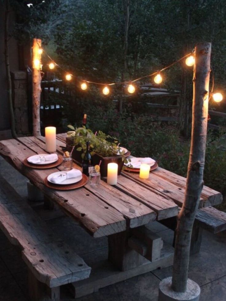 a wooden table with candles and plates on it in front of some lights hanging from the trees