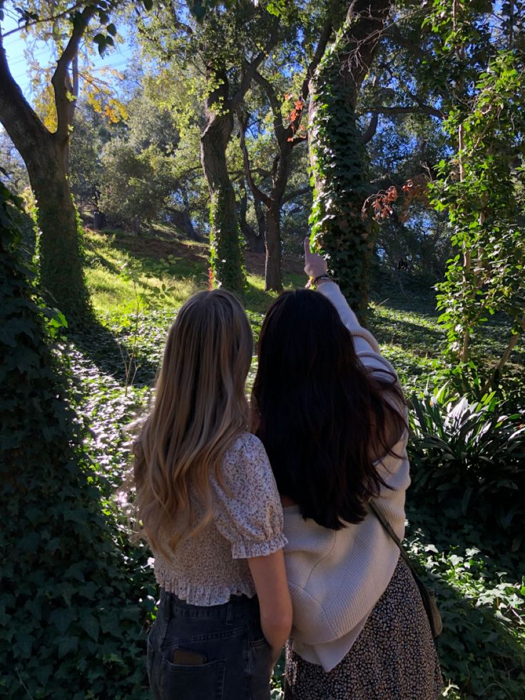 two women standing in front of trees looking at the ground with their backs to each other