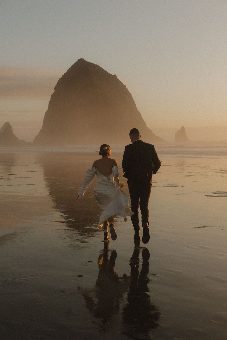 a bride and groom walking along the beach at sunset with hays in the background on a foggy day