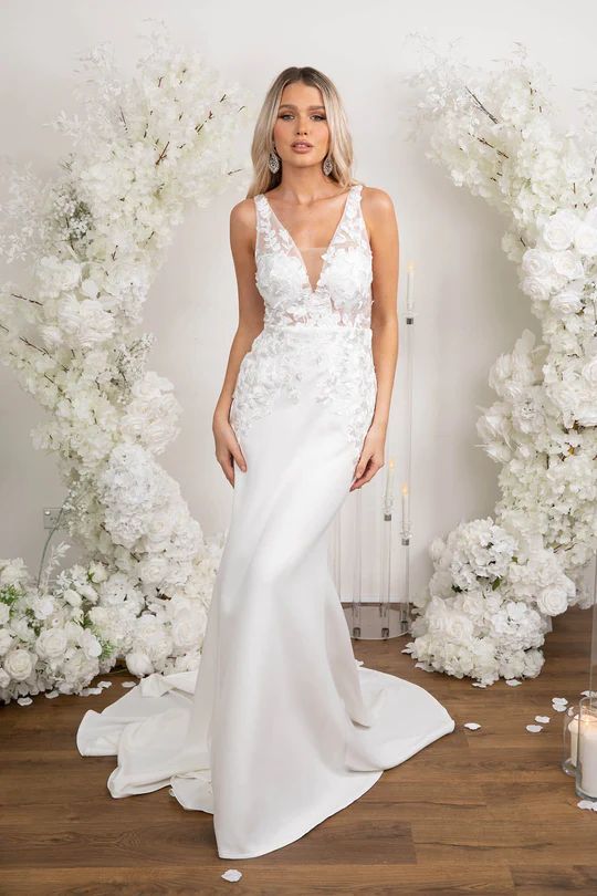 a woman standing in front of a white backdrop wearing a wedding dress with flowers on it