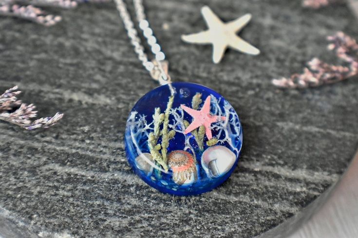 Bring a piece of the ocean with you wherever you go with our Pressed Flower "Under the Ocean" Pendant. This unique and eye-catching pendant features real blue lichen, starfish, sand, sea shells and abalone shells encased in eco-friendly resin colored with transparent inks to give it a sparkling water effect.The pendant comes with a  -1.5mm chain, 18" long, but if you prefer a different length, you can type it in the personalization box.  The pendant is available with  -925 sterling silver or  -s Ocean-inspired Jewelry With Starfish Charm As Gift, Ocean-inspired Jewelry With Starfish Charm For Gift, Adjustable Blue Necklace With Starfish Charm, Turquoise Starfish Ocean-inspired Jewelry, Ocean-inspired Adjustable Charm Necklace Gift, Ocean-inspired Sterling Silver Beach Jewelry, Ocean-inspired Blue Strand Jewelry, Blue Ocean-inspired Jewelry For Vacation, Blue Ocean-inspired Strand Jewelry