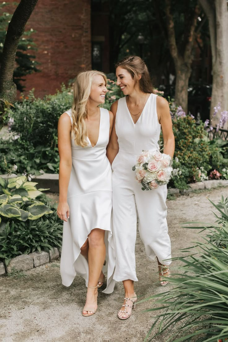 two bridesmaids in white dresses walking down the path together and smiling at each other