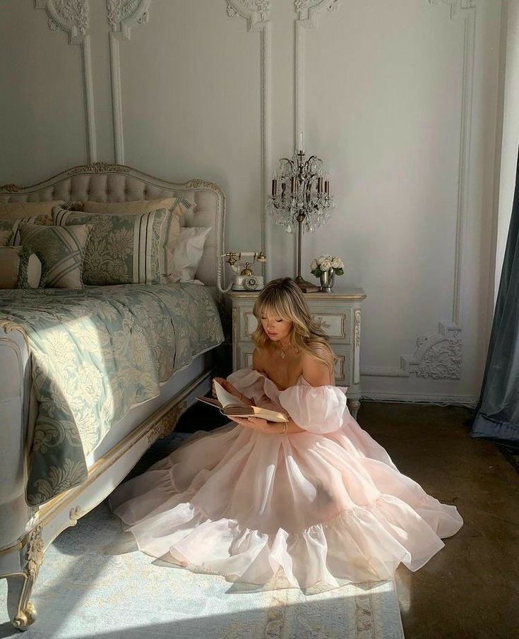 a woman in a pink dress sitting on the floor next to a bed and reading a book