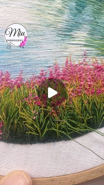 an embroidery project with flowers and grass on the side of a body of water in front of