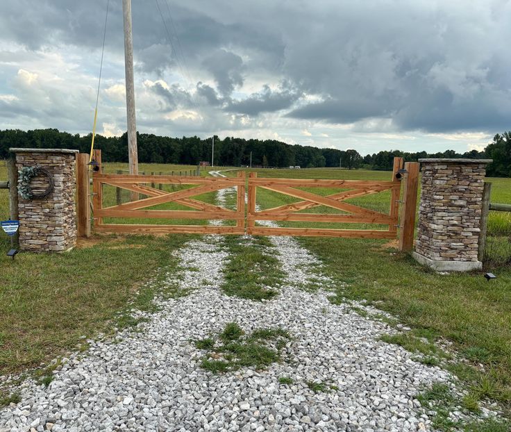 a stone and wood gate in the middle of a grassy field with gravel on the ground