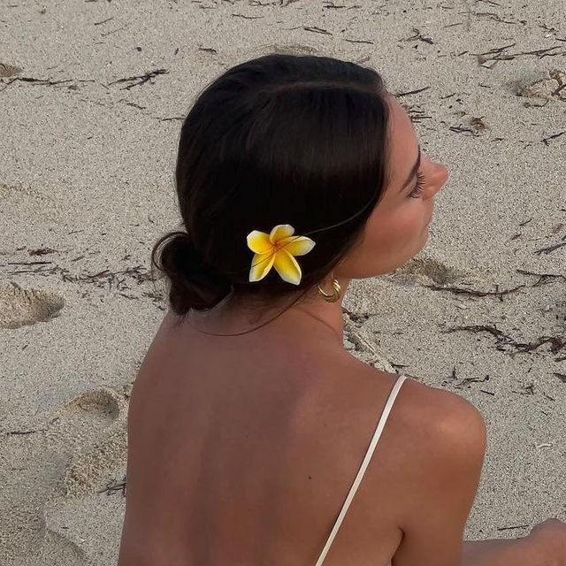 a woman sitting on top of a sandy beach next to a flower in her hair