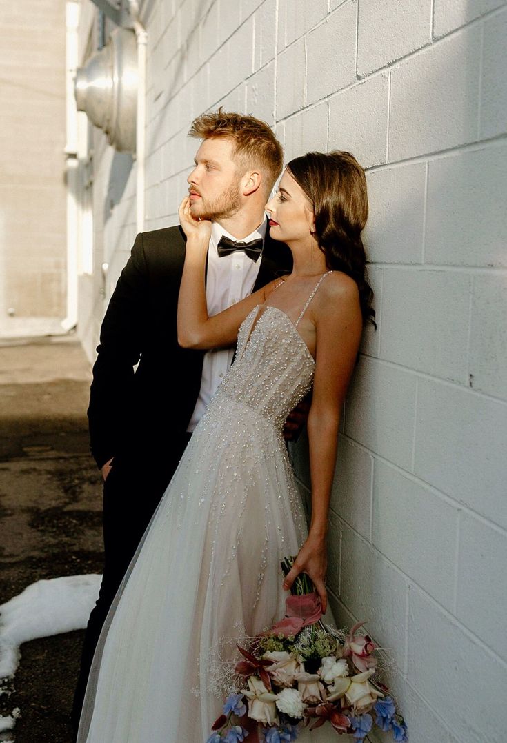 a bride and groom leaning against a white brick wall with flowers in the foreground