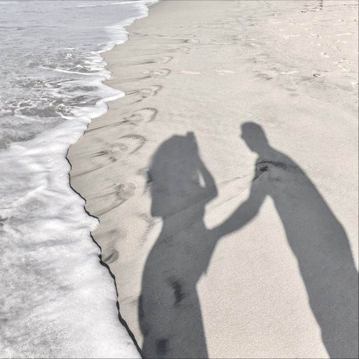 the shadow of a man and woman holding hands while standing in the sand at the beach