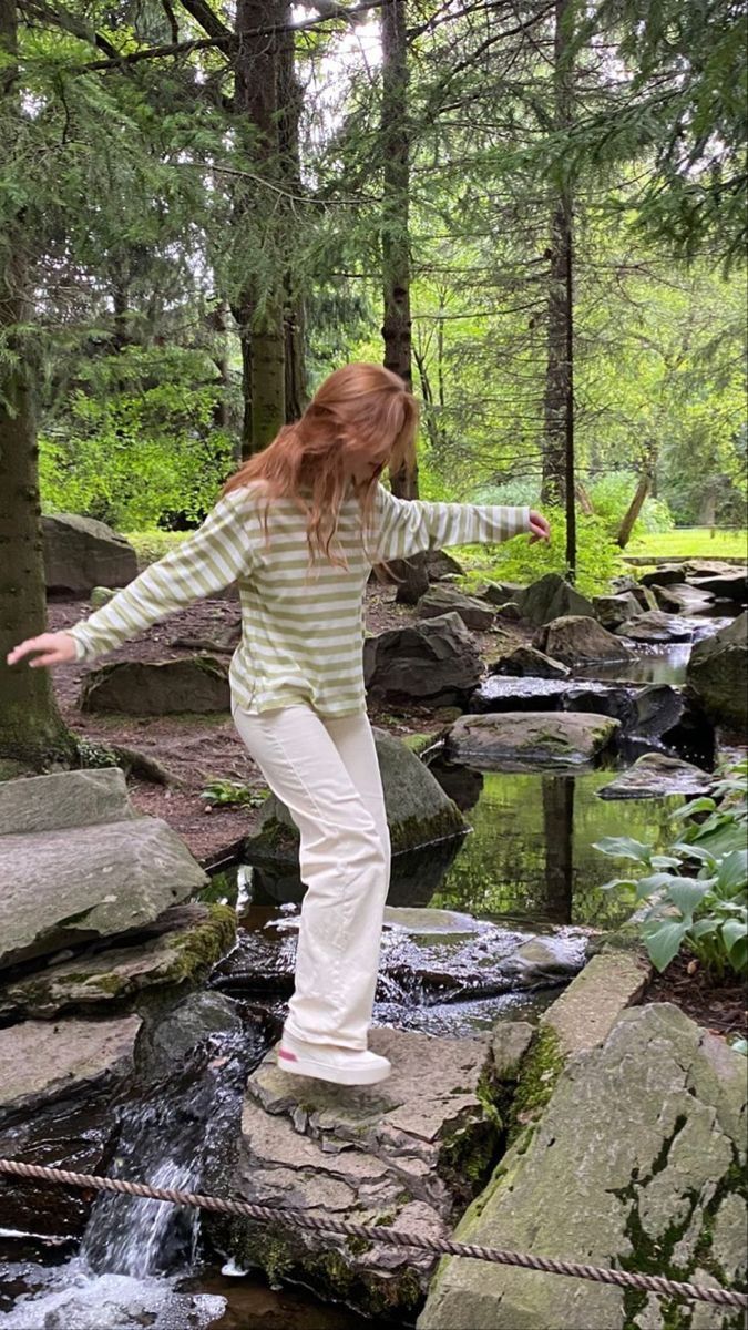 a woman standing on top of a rock next to a stream in the woods with her arms outstretched