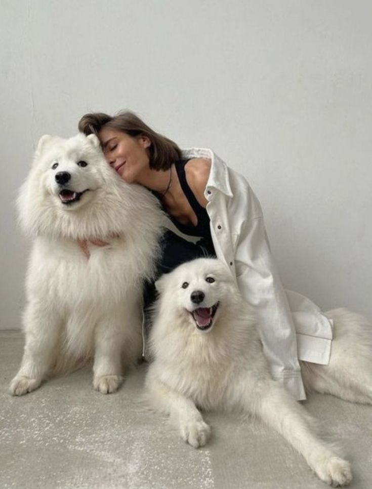 a woman sitting on the ground with two white dogs and one is hugging her head