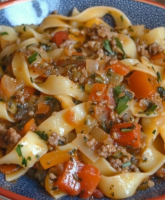 a pasta dish with sausage, carrots and other vegetables in a blue bowl on a wooden table