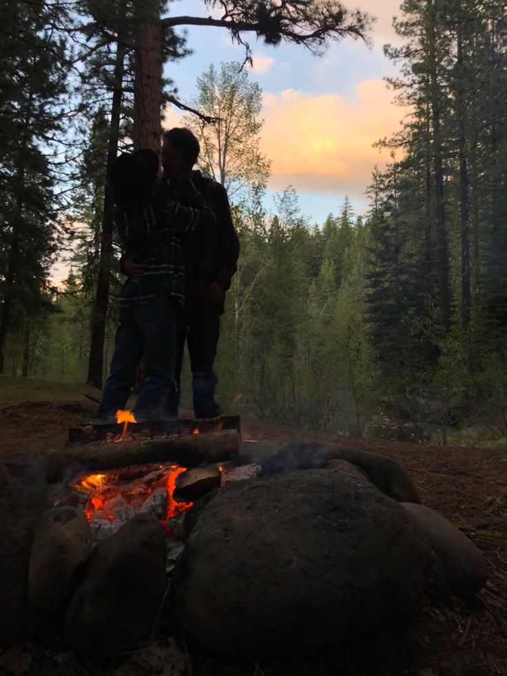 two people standing next to each other near a campfire in the middle of a forest