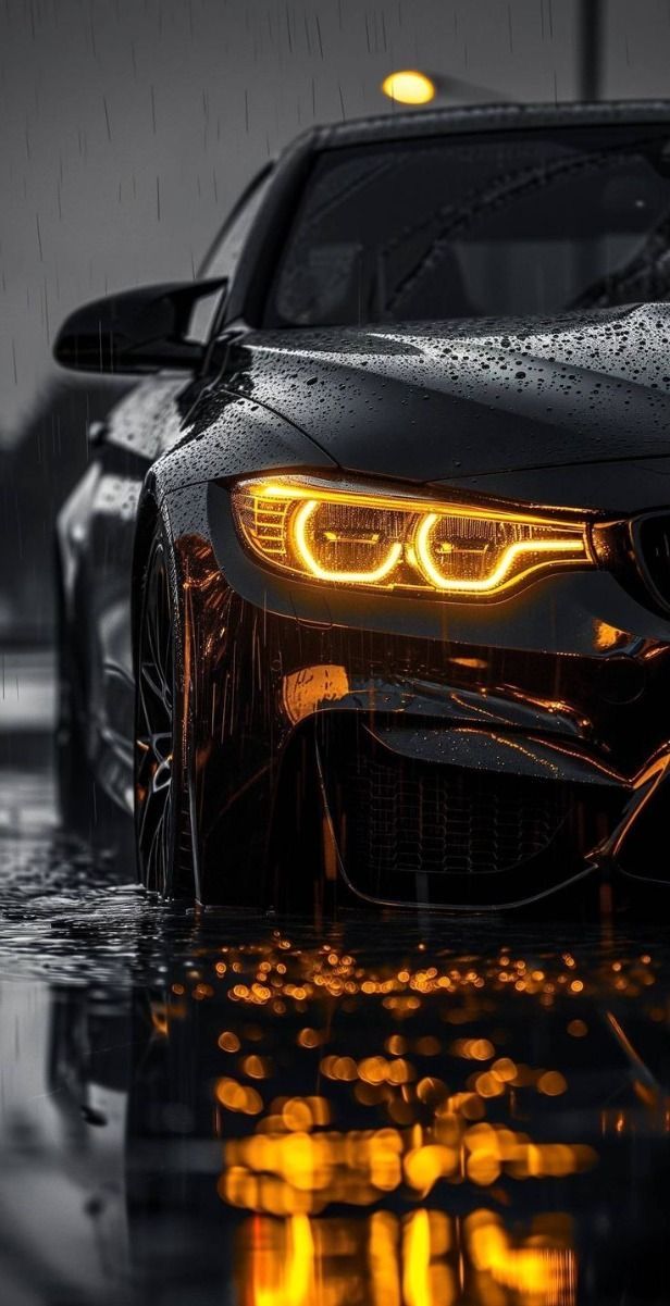 the front end of a black car with its lights on and raindrops all around it