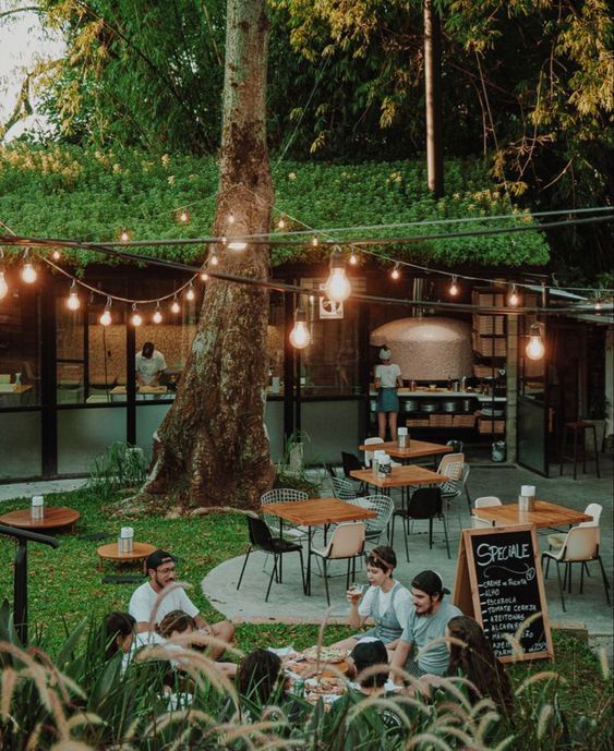 people are sitting at tables outside in the grass with lights strung over them and trees