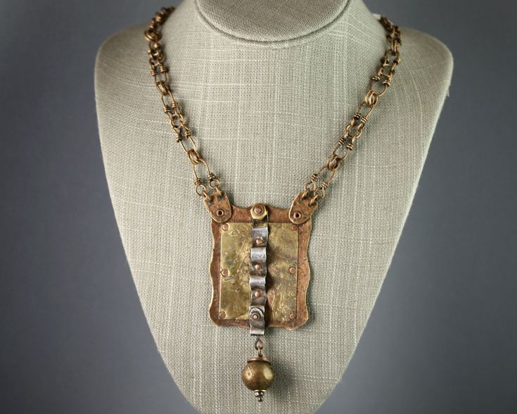 "Bronze chain necklace with large pendant composed of layered metals connected by rivets: Hammered Bronze, etched Brass, and hammered and formed Fine Silver.  A large Brass bead hangs from the bottom of the pendant. Chain is handmade with bronze wire. Dimensions of Pendant: Width at widest point 2\" (51 mm), length (including dangling bead) 4\" (102 mm). Total Necklace length:  21 3/4\" (552 mm)" Cheap Bronze Necklaces With Antique Finish, Hammered Bronze Metal Jewelry, Silver Hammered Copper Necklace, Bronze Metal Necklace With Antique Finish, Antique Finish Metal Amulet Necklace, Artisan Antique Gold Brass Necklace, Gold Amulet Necklace With Antique Finish, Bronze Antique Finish Metal Necklace, Hammered Brass Pendant Necklaces